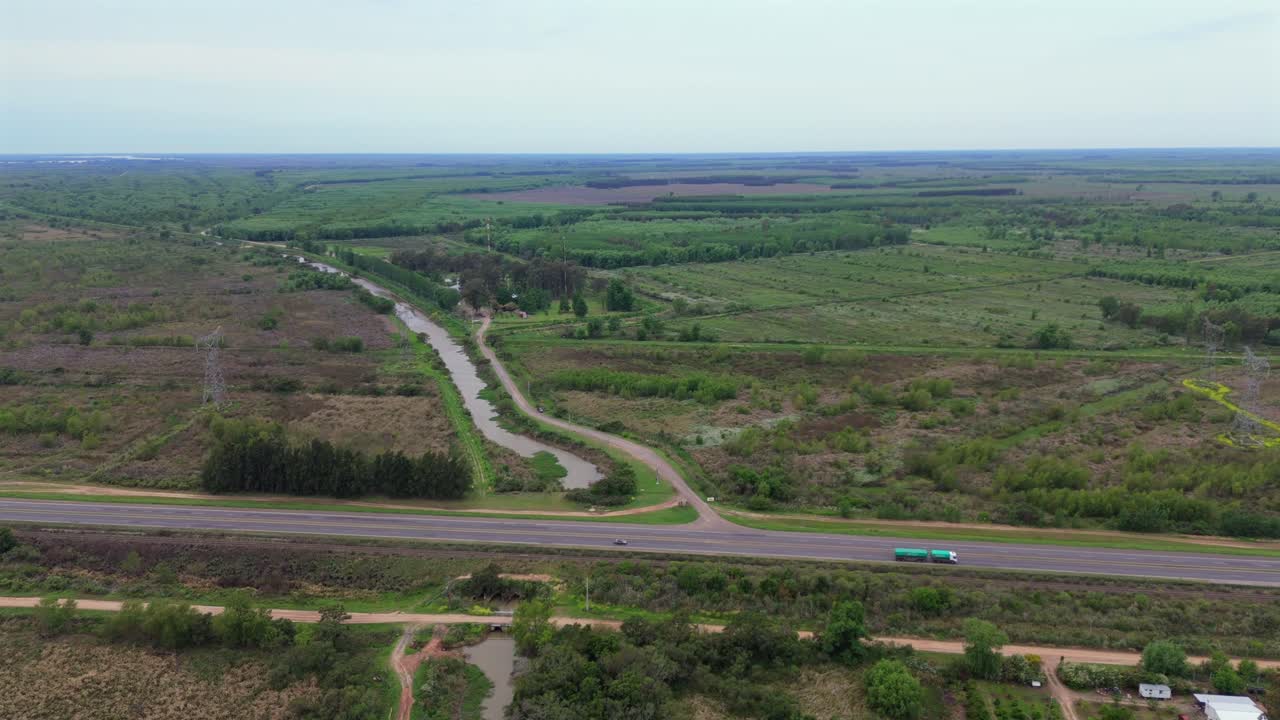 Wide drone shot of a rural landscape in the Paraná Delta, showing a highway with a passing truck, a canal, and extensive tree farms