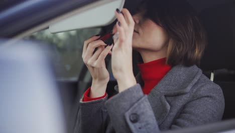 business woman in grey coat and red turtleneck applying makeup in the car