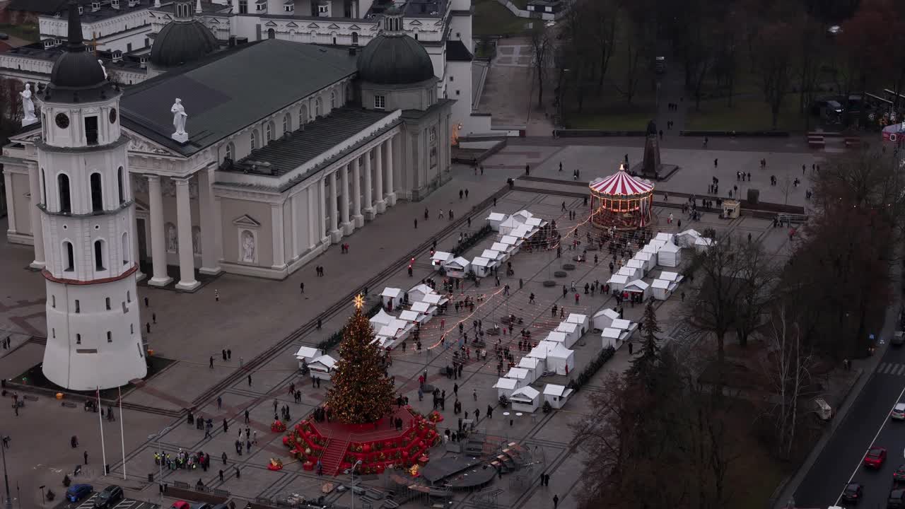 Aerial view of the Vilnius Christmas Market in Lithuania during a cloudy winter day showing a large christmas tree and many vendor tents with the Place of the Grand Duke of Lithuania in the background