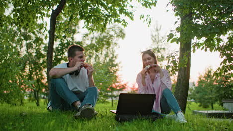 brother and sister seated outdoors with laptop in front making a toast with bread in hand, enjoying meal together in natural sunlight filtering through trees