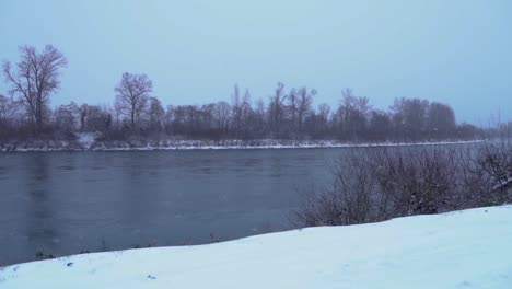 Nieve-Cayendo-En-Tormenta-Junto-Al-Río,-Cámara-Lenta,-Panorámica-Hacia-La-Izquierda