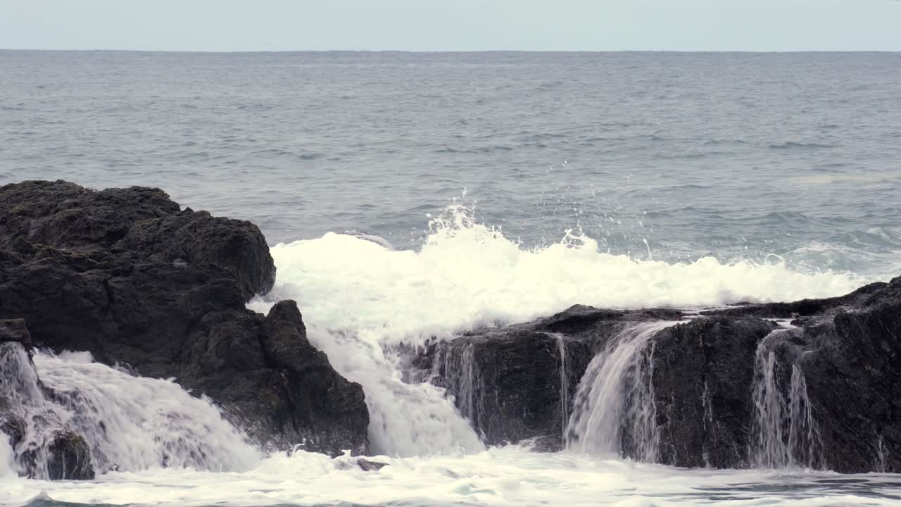 Waves crash against dark, rugged rocks on a serene coastline, creating white foam under a cloudy sky. The ocean stretches to the horizon, evoking tranquility