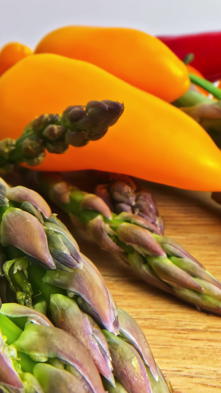 Macro pull-back close-up of fresh asparagus spears with colorful orange sweet peppers and red chili on wooden board