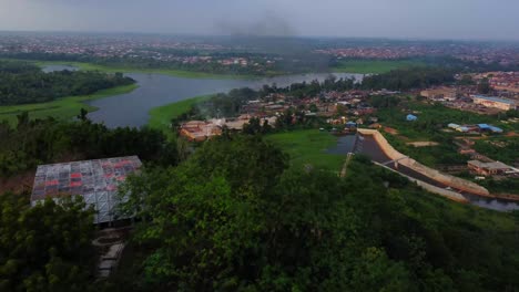 drone flying over a mountain filled with green trees and revealing a polluting industrial zone with black smoke alongside eleyele river in nigeria, africa