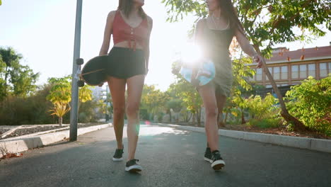 Entre-Palmeras,-Las-Amigas-Caminan-Hacia-El-Parque,-Cargando-Patinetas,-Conversando,-Riendo-Y-Sonriendo-Alegremente-En-Los-Cálidos-Tonos-Del-Atardecer,-Todo-En-Cámara-Lenta.