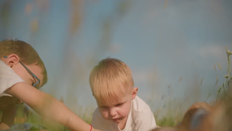 two young boys in white tops are enjoying a sunny day on a checkered picnic blanket in a grassy field. one boy is lying down while the other is bent over, both focused on eating bread together