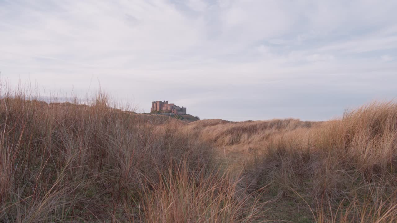toma fija a nivel del suelo del castillo de bamburgh en la distancia, con altas dunas de arena cubiertas de hierba en primer plano