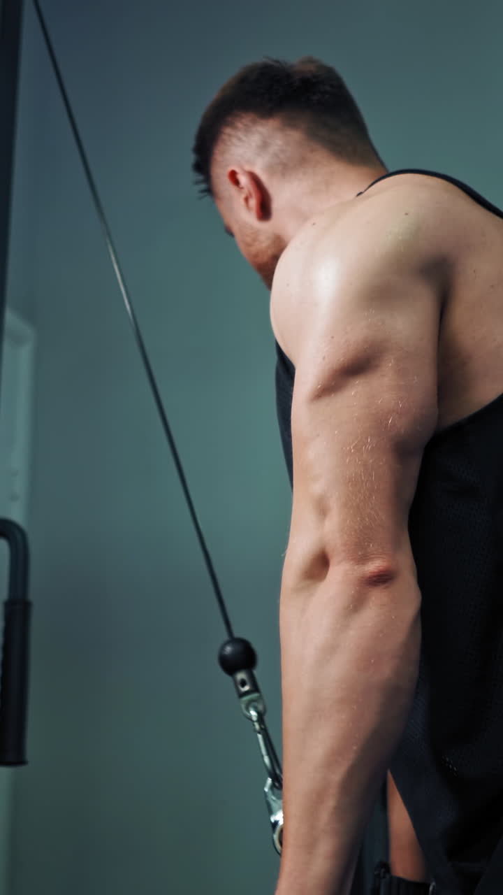 Muscular man trains on the simulator in fitness center. Strong athlete pull bar with rope and weights on fitness machine indoors. Vertical video