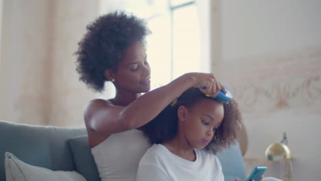 Afroamerikanische-Mutter,-Die-Das-Haar-Ihrer-Tochter-Kämmt,-Sitzt-Auf-Der-Couch,-Während-Ein-Kleines-Mädchen-Ein-Handy-Benutzt