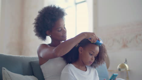 african american mum combing her daughter's hair sitting on couch while little girl using mobile phone