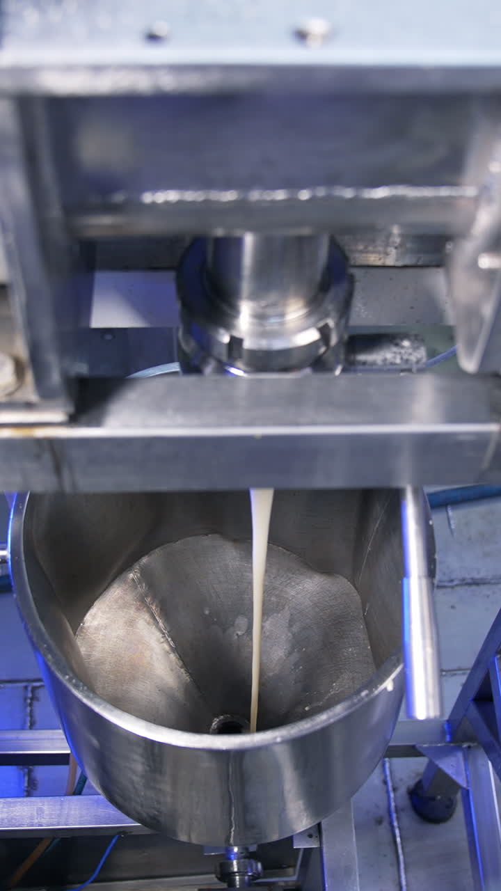 Thick creamy condensed milk being poured from a pipe into a metal tank. Confectionery production line at food factory. Vertical video
