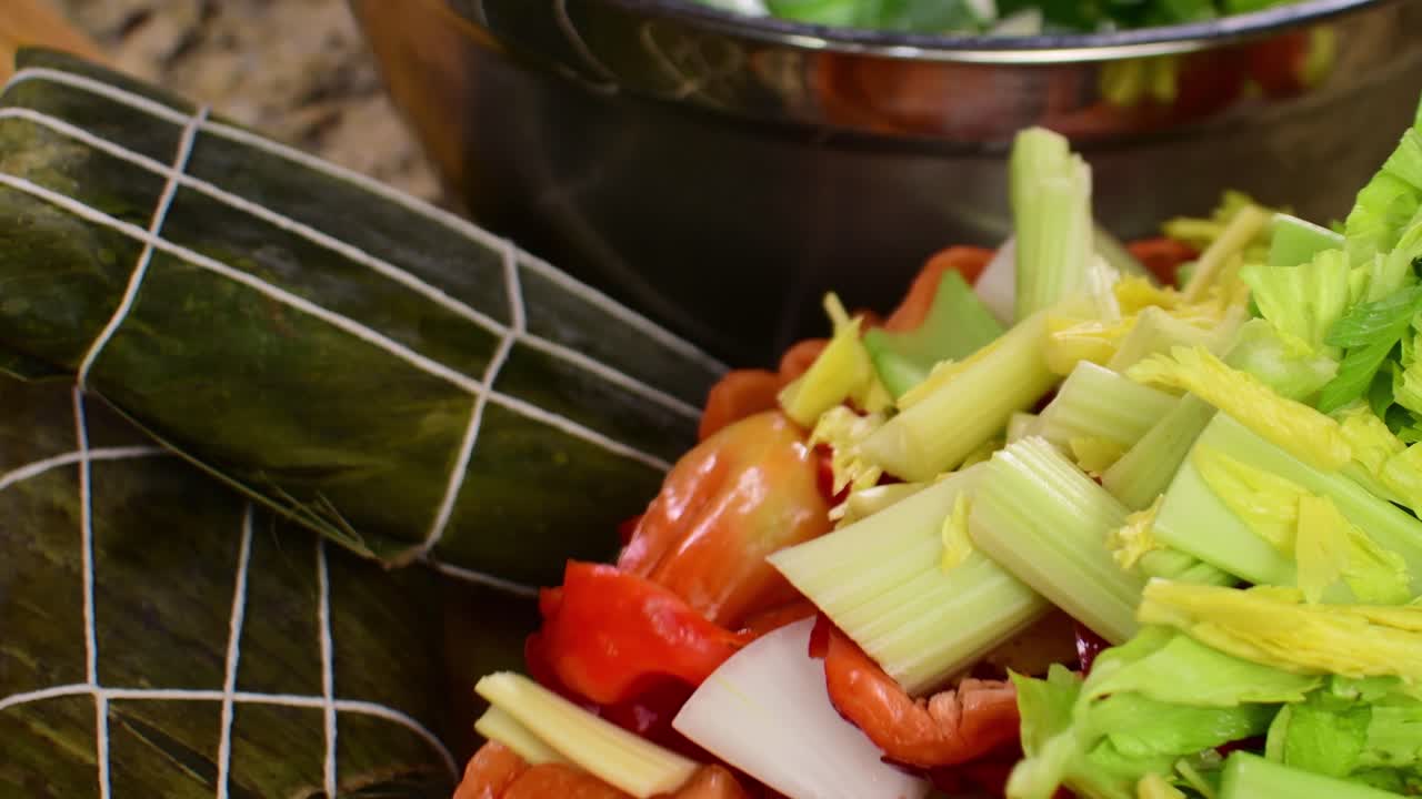 High-angle, close-up shot of raw hallacas wrapped in banana leaves next to colorful, fresh vegetables spinning on a turntable