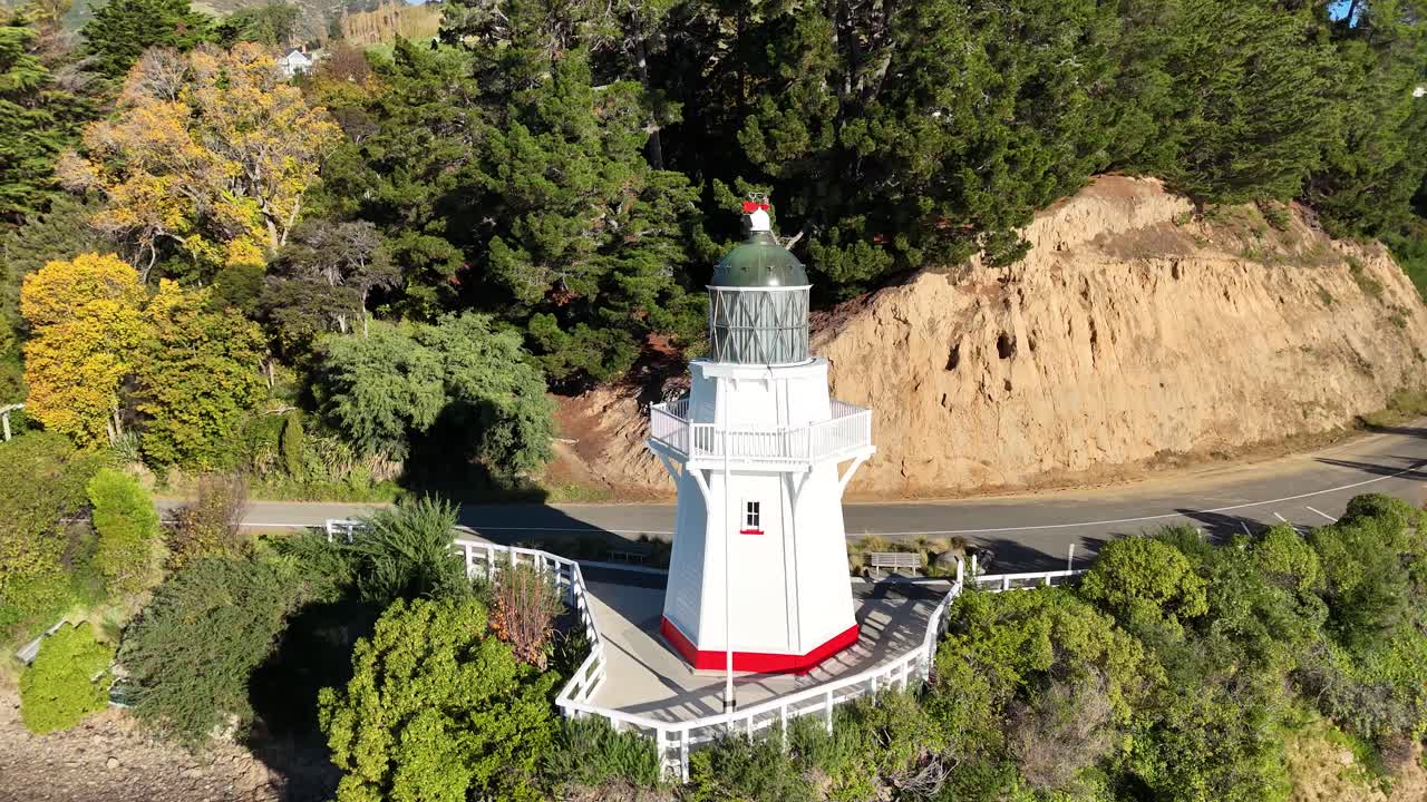 Drone footage captures a scenic lighthouse on a cliff surrounded by lush greenery in Akaroa, New Zealand, under bright daylight