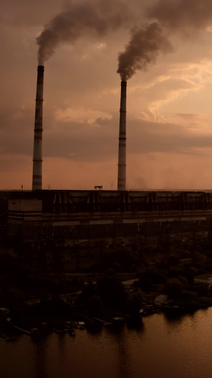 Factory with smoke pipes in evening nature. Dangerous manufacturing pollute atmosphere. Industrial plant near the river at dusk. Ecological problems. Vertical video