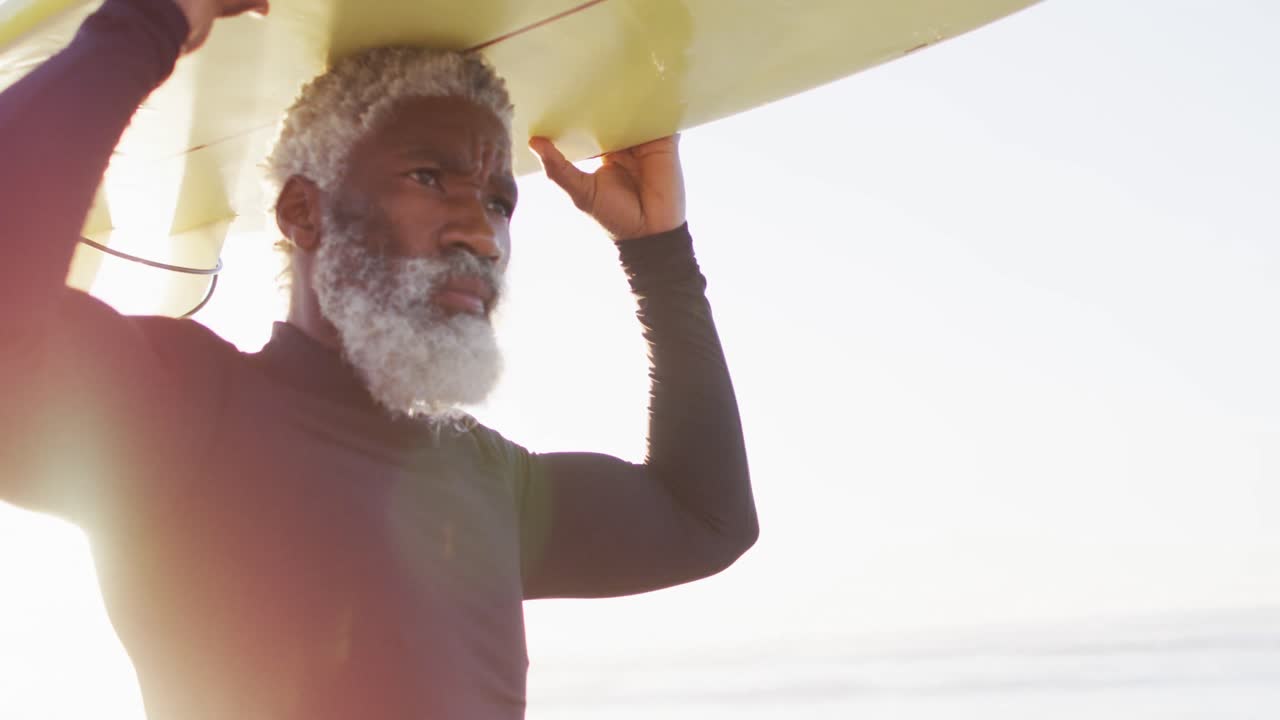 hombre afroamericano mayor caminando con una tabla de surf en una playa soleada