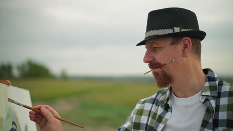 close-up of a man wearing a hat and checkered shirt, intensely focused on painting. he holds a brush in his mouth while working on his artwork, capturing the essence of creativity and concentration