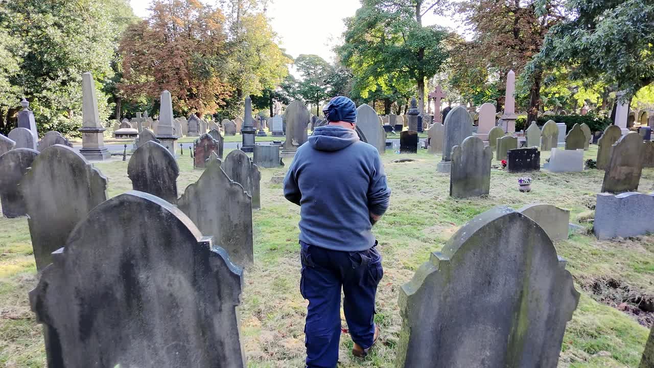 Following behind male wearing hat and casual clothing walking alone in peaceful sunlit cemetery