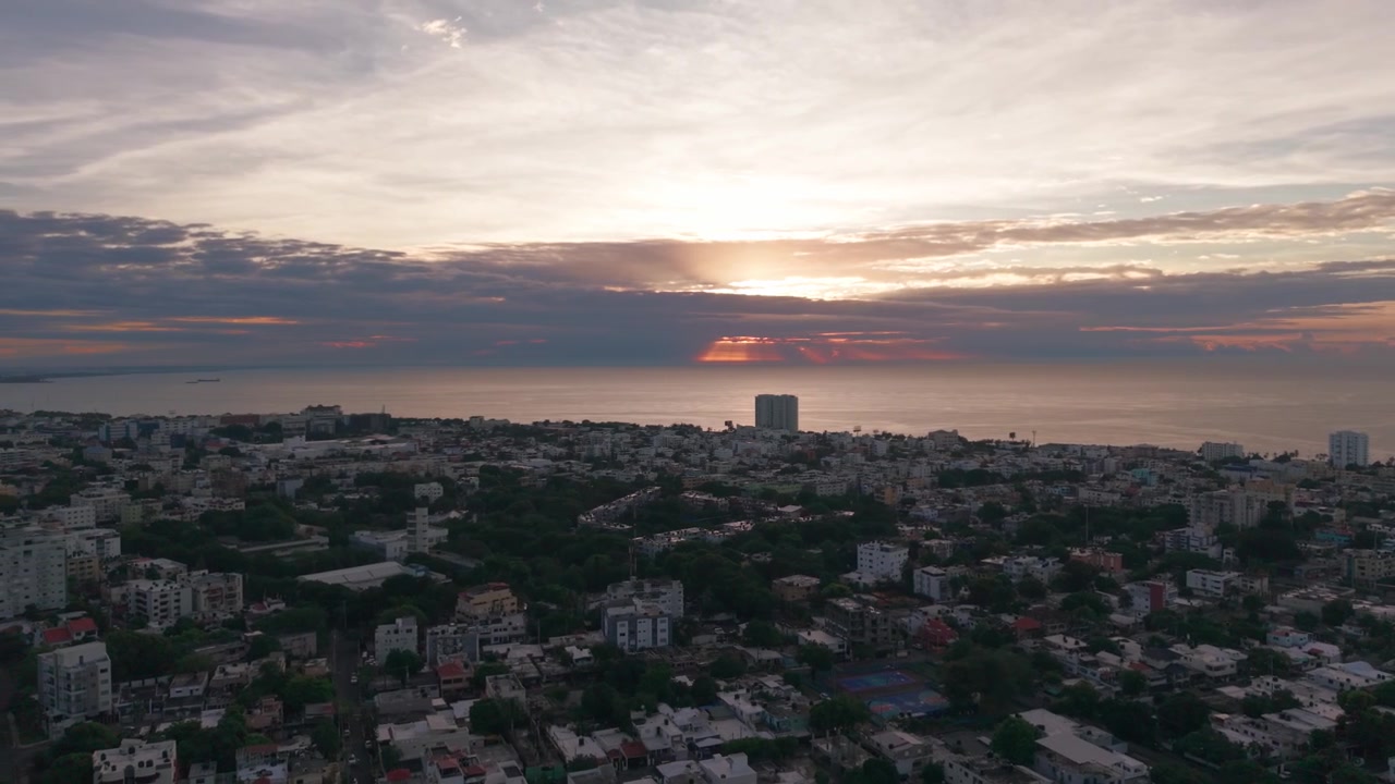 Hochwinkel-Luftbild-Drohnen-Schwenk nach links über der Stadtlandschaft von Santo Domingo, der einen dramatischen bewölkten Sonnenaufgang über dem Karibischen Meer und dem grünen Blätterdach des Mirador Sur Parks enthüllt.