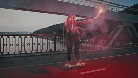 joven mujer hipster en traje informal está montando patineta en el puente sosteniendo una llamarada de señal roja en llamas y agitando