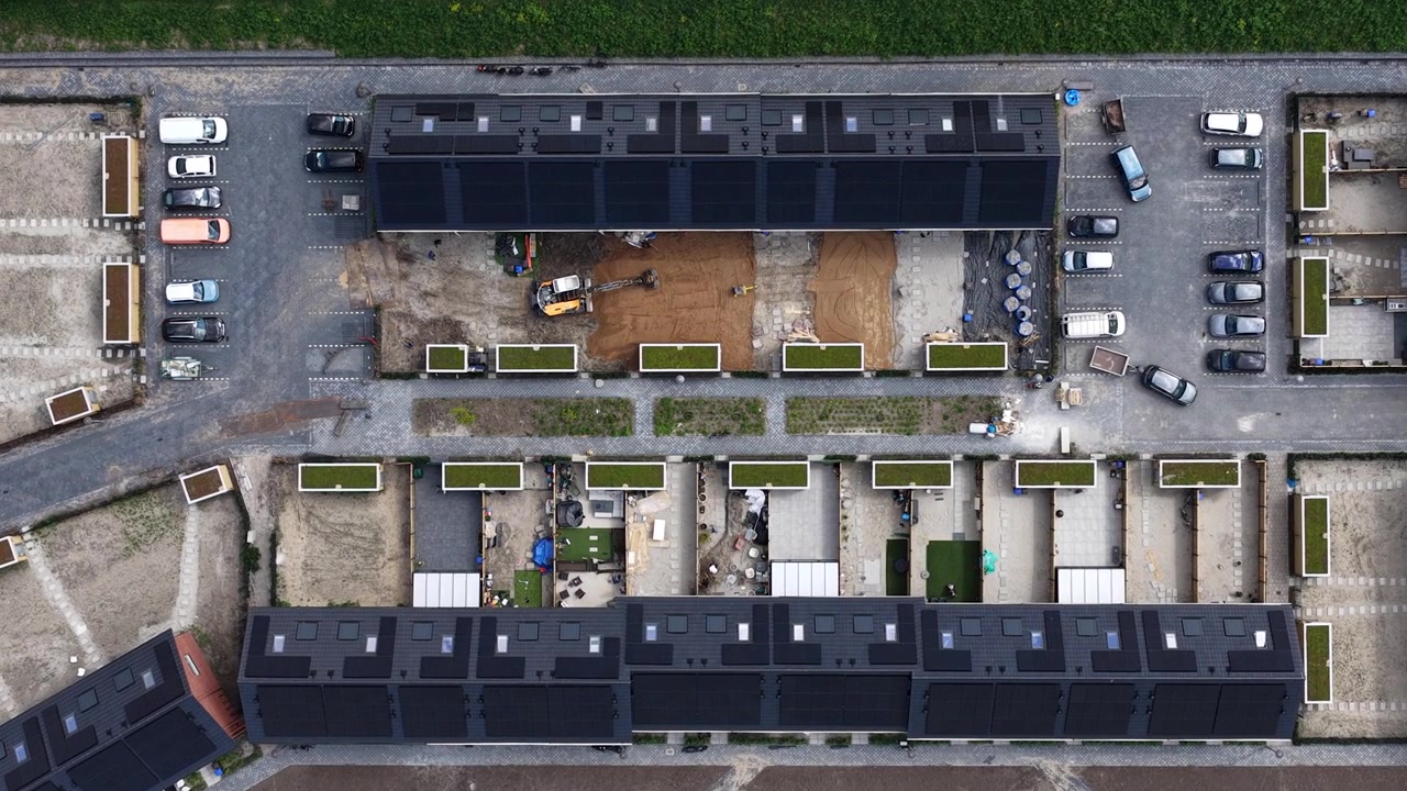 Vista aerea dall'alto con angolazione elevata che mostra moderne case a schiera in costruzione, con tetti dotati di pannelli solari e tetti verdi, tra lavori di paesaggistica e preparazione del terreno in corso in un nuovo complesso residenziale.