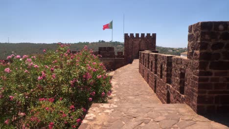 Dentro-De-Los-Muros-Del-Castillo-De-Silves,-Portugal-Con-Adelfa-Rosa-En-Primer-Plano-Y-La-Bandera-De-Portugal-Ondeando-Al-Fondo-Bajo-Un-Cielo-Azul-Brillante