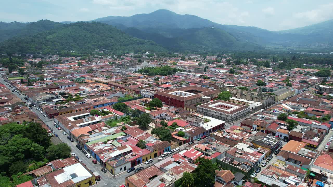 Riprese aeree con drone della città coloniale di Antigua, Guatemala, che mostrano tetti rossi vivaci e colorati e cime di alberi verdi, circondata da lussureggianti colline e montagne di giungla