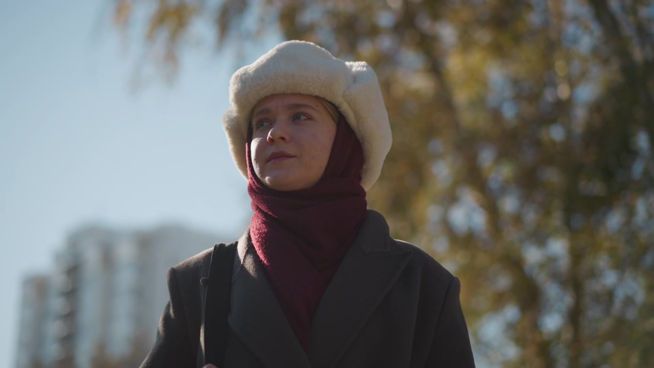 Primo piano di una donna che indossa abiti invernali all'aperto con edifici e alberi sullo sfondo. La donna è vestita con un cappotto scuro, una sciarpa bordeaux e un cappello bianco mentre tiene un oggetto in mano. La scena