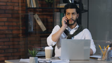 Elegante-Hombre-De-Negocios-árabe-Sentado-En-Su-Escritorio-Con-Una-Laptop-En-La-Oficina-Y-Hablando-Por-Teléfono,-Luego-Se-Levanta-Y-Se-Va