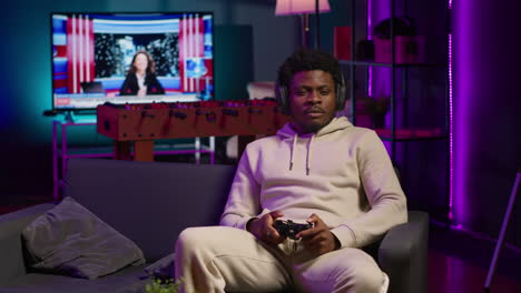 cheerful man in his apartment playing video games on couch, enjoying day off from work. black gamer battling enemies in online multiplayer shooter on gaming console. camera a.