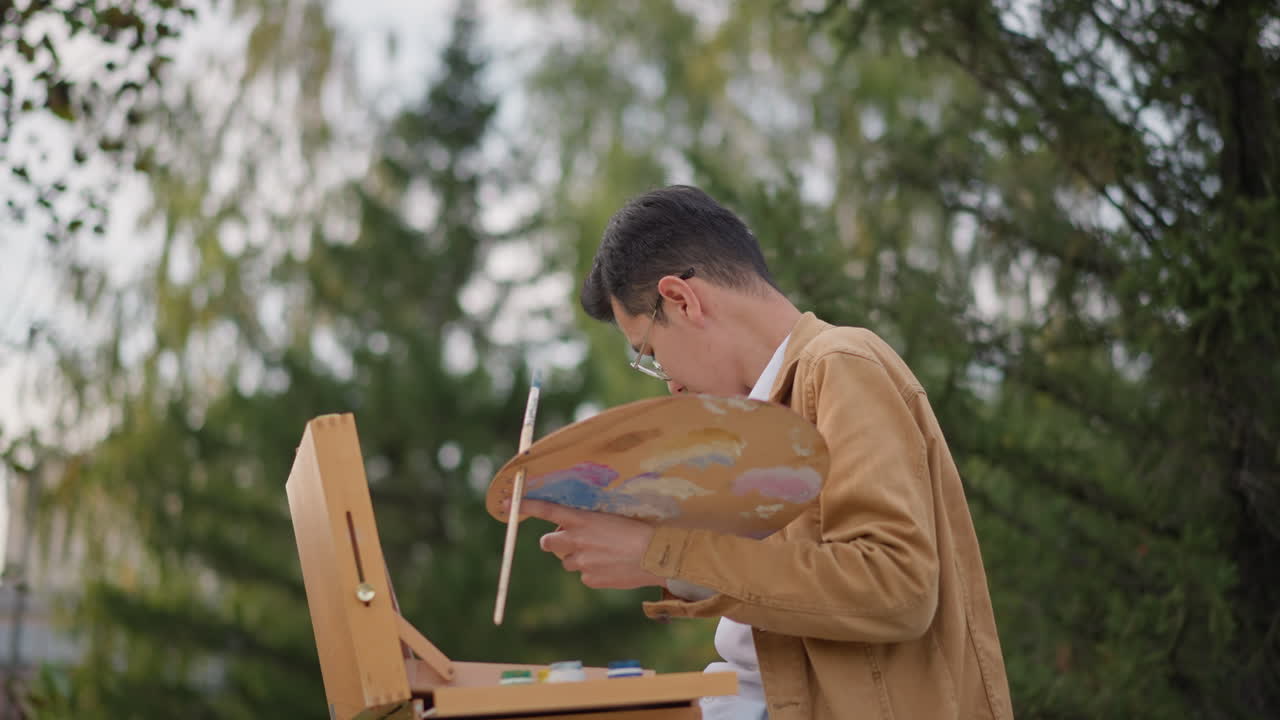 パレットとイーゼルを持って屋外で絵を描く若いアジア人アーティスト、油絵の具を混ぜる茶色のジャケットを着た男性、キャンバスに風景のスケッチ、緑豊かな公園の背景、柔らかい自然光、穏やか。