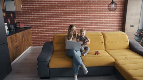 father plays with his son while their mother busy with her laptop on the sofa