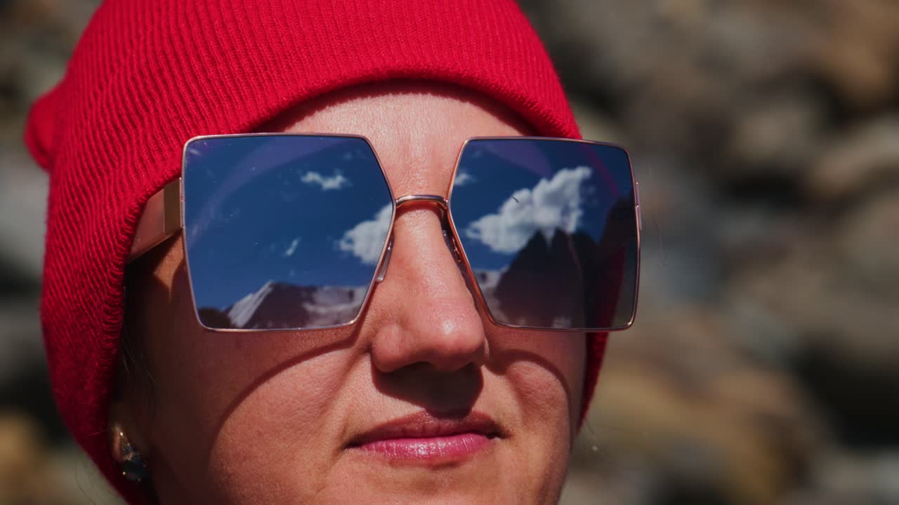 Kvinne med rød lue og reflekterende solbriller utendørs. Solbrillene speiler fjelltopper og blå himmel. Hun ser rolig ut, fokusert på omgivelsene. Ideell for eventyr og reise.