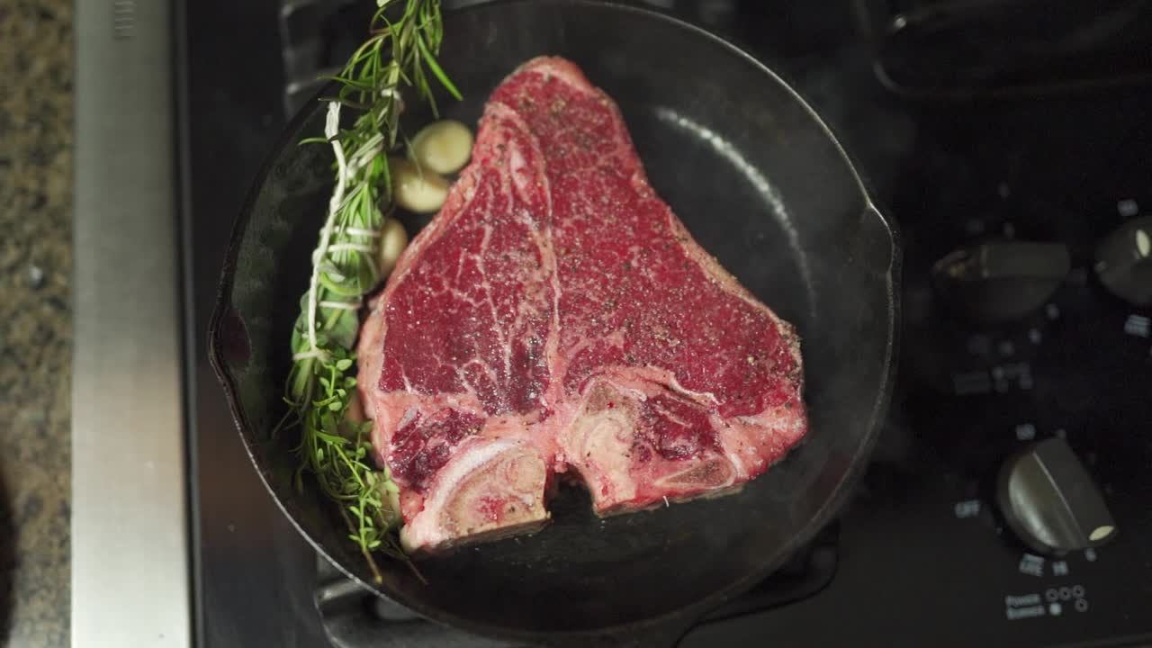 Slo-mo footage moving over a juicy steak, garnished in herbs and spices, slowly cooking in a cast iron pan on top of an oven range.
