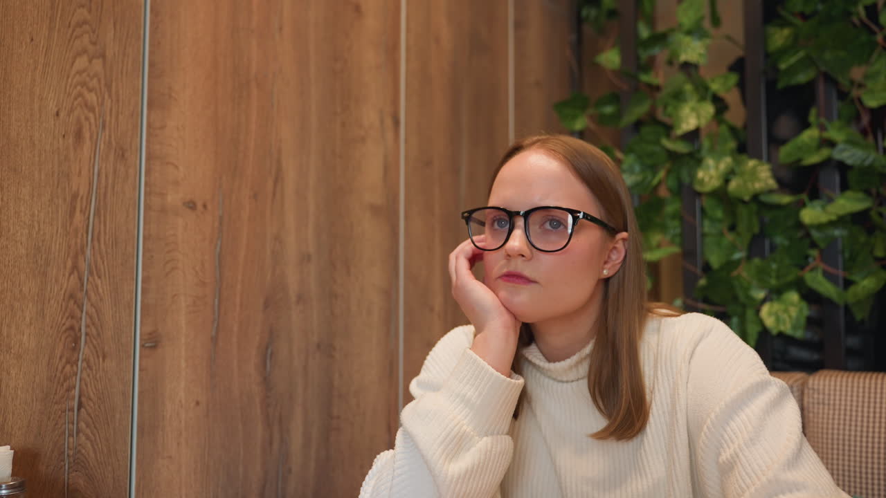 Student in glasses and white sweater sits indoors resting head on hand while gently tapping cheek in thoughtful mood, wooden wall background and container with items visible on left side