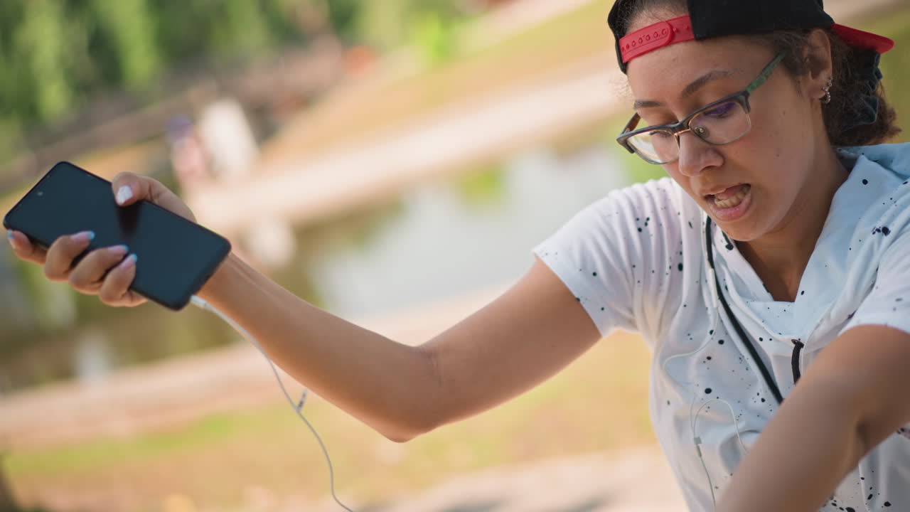 Giovane che balla in un parco con cappellino all'indietro e occhiali, ascoltando la musica con le cuffie con filo da uno smartphone, gesticolando in modo espressivo con le braccia e con un'espressione concentrata vicino al sentiero del laghetto in una giornata di sole