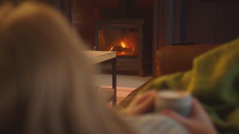 Close-Up-Of-Woman-At-Home-In-Lounge-Lying-On-Sofa-Looking-At-Cosy-Fire-With-Hot-Drink