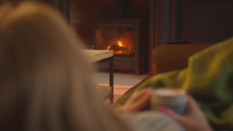 cerca de la mujer en casa en el salón acostado en el sofá mirando el fuego acogedor con bebida caliente