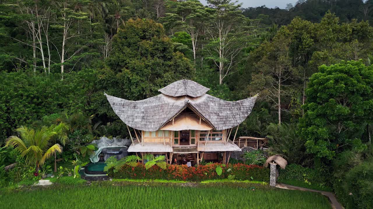 Aerial drone footage of a tropical bamboo house with a small pool, surrounded by lush rice fields and dense forest in the background, showcasing Bali’s serene nature, eco design, and peaceful scenery
