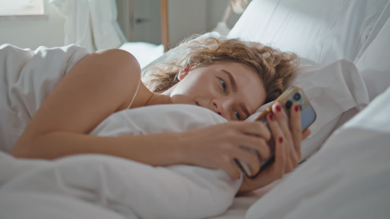 Nærbillede af fredelig kvinde, der sover i sengen. Søvnig krølhåret pige tjekker smartphone.