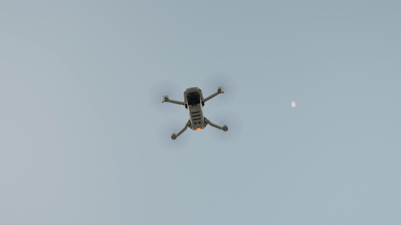 A drone hovers high in the sky during a clear day with the moon visible in the background