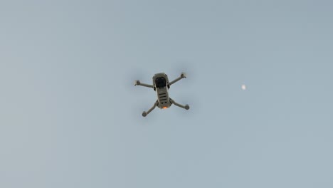 a drone hovers high in the sky during a clear day with the moon visible in the background