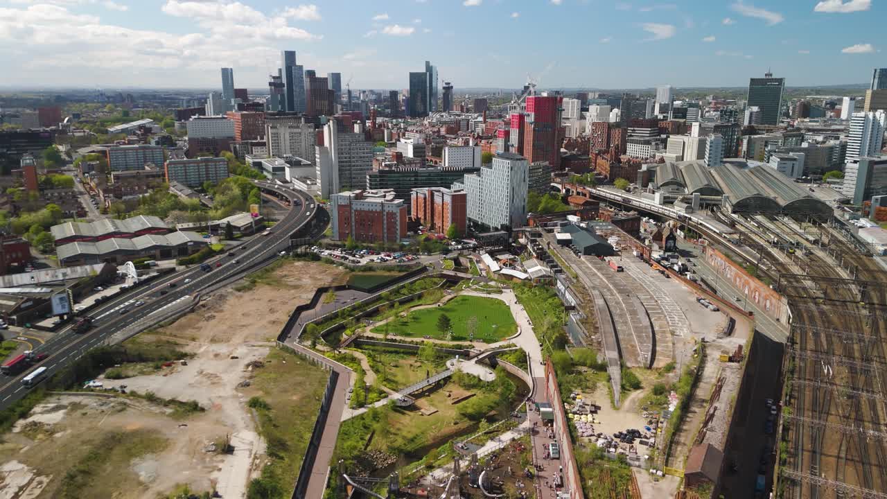 Plano aéreo em movimento de drone do centro da cidade de Manchester, com estação de trem e carros ao longo da via principal. Céu azul com nuvens fofas e parque verde abaixo
