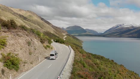 Motorhome-driving-along-the-coast-of-Lake-Wakatipu,-Queenstown,-New-Zealand-with-mountains-fresh-snow-in-background---Aerial-Drone