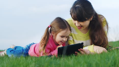 una madre joven y bonita jugando en una tableta junto con su pequeña hija en un claro verde