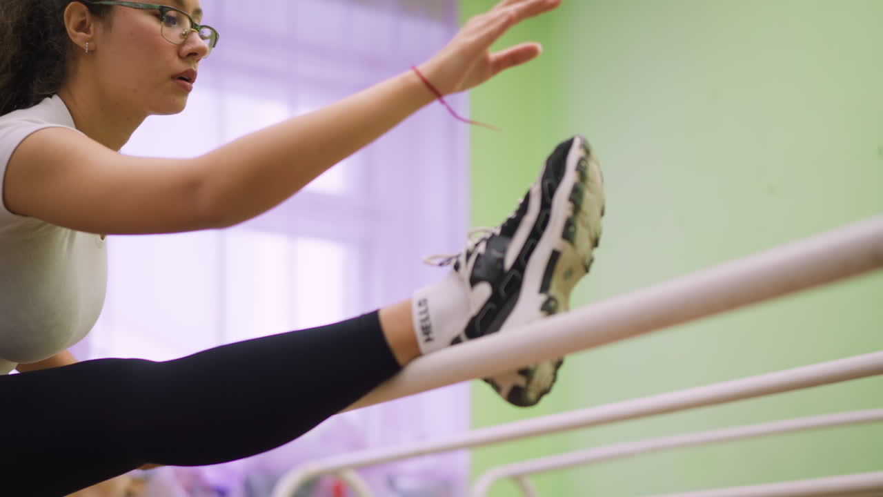 Girl in white top and black leggings places leg on bar while stretching gently indoors, wearing black sneakers and white socks, extending arm forward with focus during fitness flexibility routine