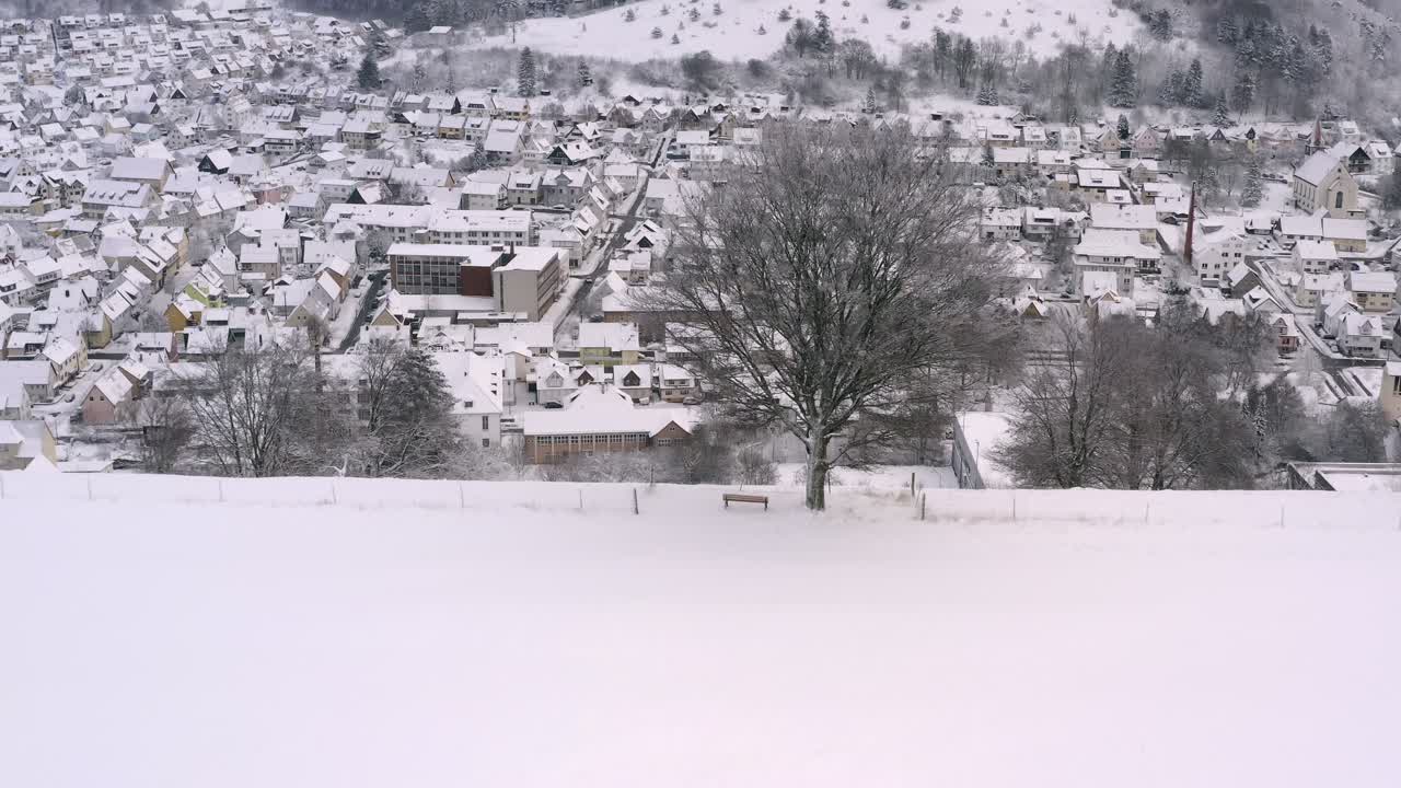드로니 샷, 벤치가 있는 눈 덮힌 나무에서 뒤로 벗겨져 눈 덮힌 하얗고 꿈결 같은 마을이 있는 계곡을 발견합니다.