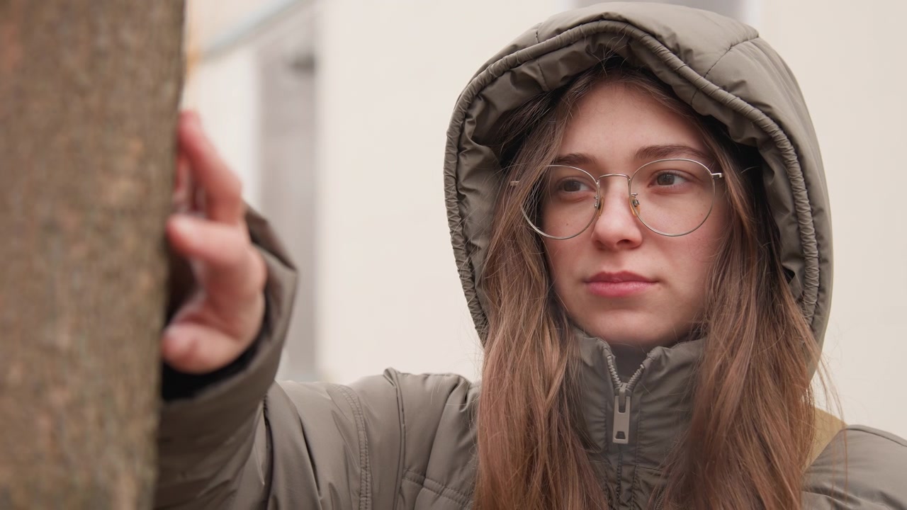 Donna caucasica con cappotto con cappuccio in piedi accanto a un albero, mano che tocca la corteccia, occhiali, capelli lunghi castani, sfondo urbano neutro, leggera neve sul cappuccio, espressione contemplativa, toni smorzati, freddo.