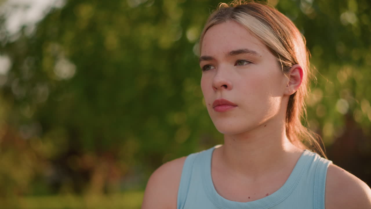 집중된 표정을 가진 젊은 여자의 클로즈업, 롤러 스케이팅처럼 외부에서 우아하게 움직이고, 빛이 그녀의 얼굴에서 반사되고 머리카락이 뒤로 여, 흐릿한 배경에 울창한 나무로 둘러싸여 있습니다.