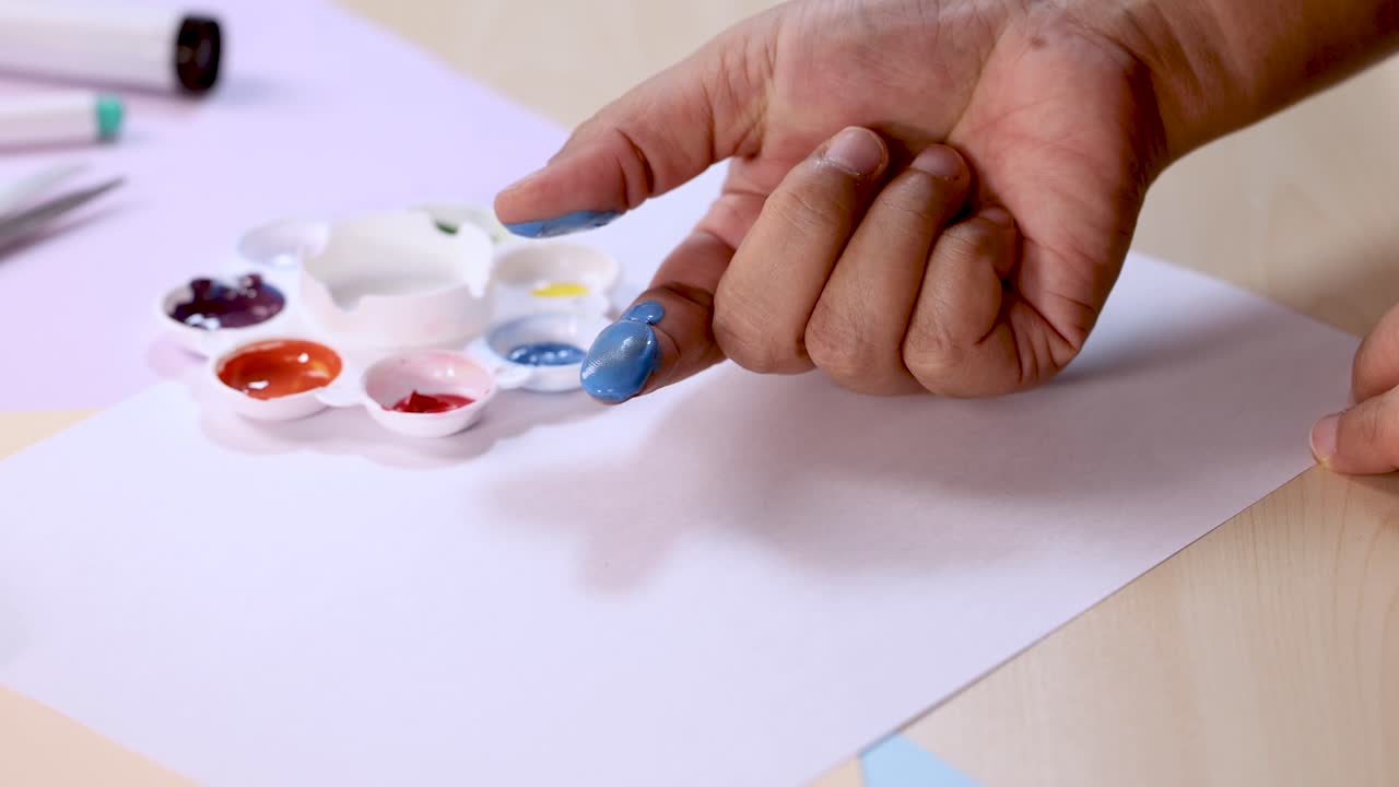 Hand dips finger in blue paint, preparing for fingerprint art on white paper, bright lighting