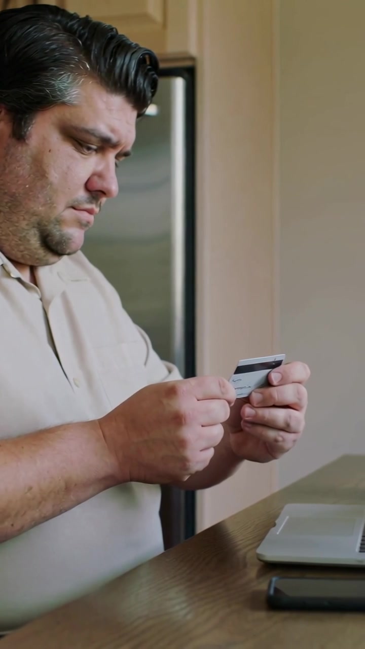 Video verticale: Uomo in polo beige che ispeziona una carta di credito e digita il pagamento sul laptop al tavolo della cucina.